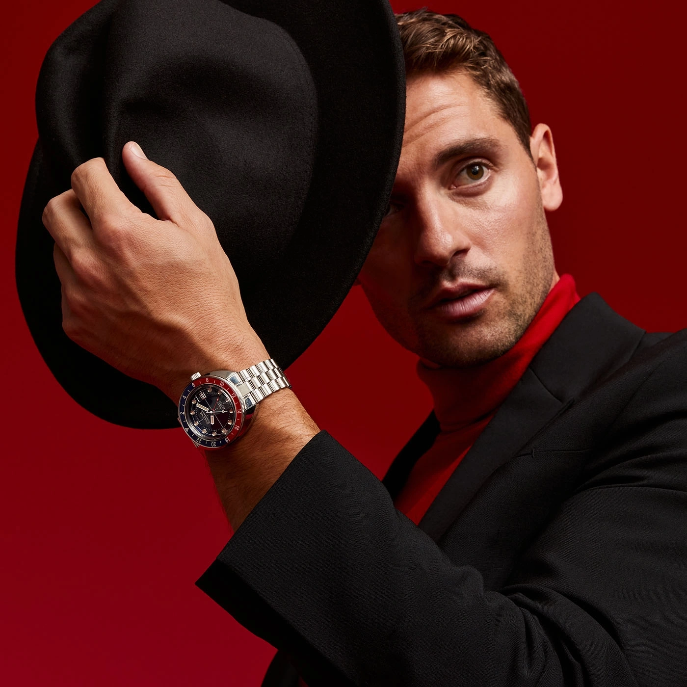 Mann mit Hut und Uhr, roter Hintergrund, schwarzer Anzug, roter Rollkragenpullover