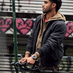 Mann mit Fahrrad und Uhr, vor Graffiti-Hintergrund, lässig gekleidet.