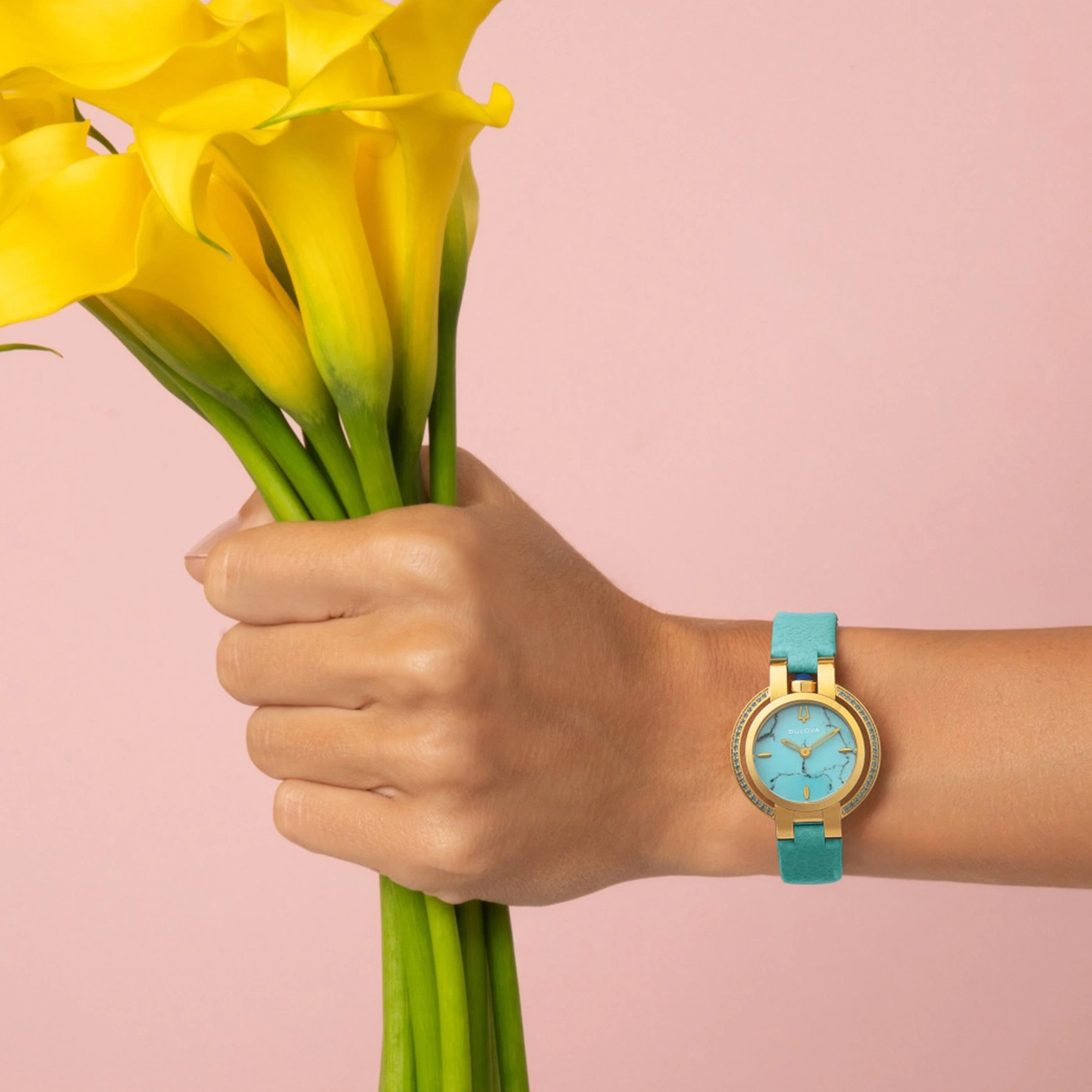 Eine Hand hält gelbe Callas, trägt eine goldene Uhr mit türkisfarbenem Zifferblatt und Band.